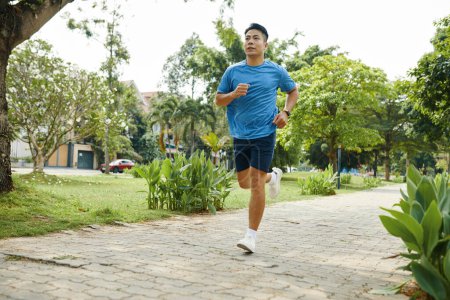 Hombre joven con una camisa azul corriendo a lo largo de un sendero del parque durante su rutina de ejercicios matutinos