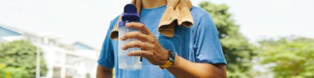 Close-up of a man holding a water bottle with a towel draped over his shoulders after a run in the park, focusing on hydration and recovery