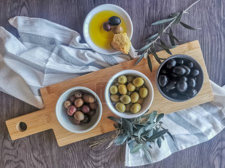 Photo for View at the different types of olives inside bowls on top of a traditional wooden base, plate with extra virgin olive oil and a piece of cornbread, dark wood table background and olive branches... - Royalty Free Image