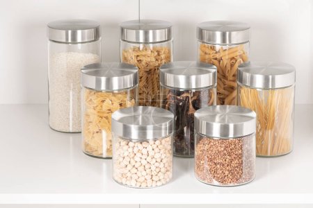 Téléchargez les photos : Un gros plan d'une variété d'assaisonnements et de céréales stockés dans des pots en verre transparent et des conteneurs de stockage sur une étagère en bois blanc - en image libre de droit