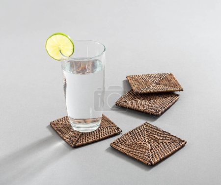 Glass of water with lemon beside wicker coasters set on grey background.