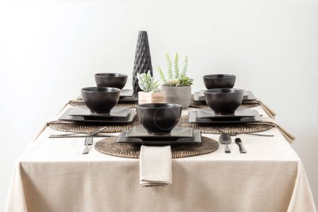 Photo for Table with beige tablecloth setting with black plates, silverware and succulent plant ornament, inside a dining room - Royalty Free Image