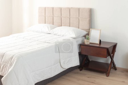 Interieur eines modernen Schlafzimmers mit einem weißen Bett mit einem bequemen Laken und einem hölzernen Nachttisch sowie einem beigen Kopfteil aus Stoff