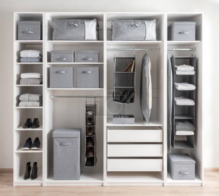 Weiße Garderobe mit verschiedenen Kleidungsstücken und graue Abstellbehälter für Accessoires. Der Kleiderschrank verfügt über weiße Holzregale