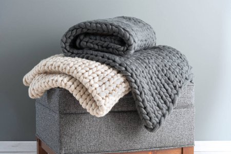 Folded knitted plaid blankets on a stool against a gray wall, close-up.