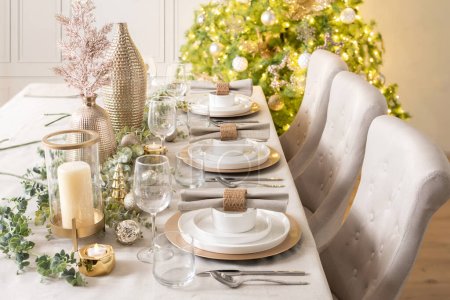 Tischgedeck für das Weihnachtsessen, Geschirr auf goldenen Ladetellern in weiß und goldenen Farben mit Kerzen, in einem skandinavischen Haus Interieur