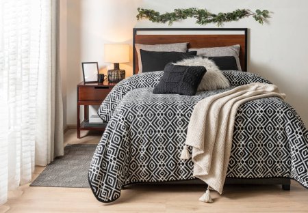 Interior of a modern Nordic bedroom with a comfortable bed featuring a geometric blanket, black and beige pillows on the bed, and a wooden bedside table