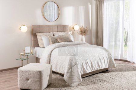 Photo for Modern Scandinavian-Style Bedroom with Neutral Tones, Featuring a Double Bed with White Cover, Rectangular Pouf, and Mirror, Bathed in Neutral Daylight from a Window - Royalty Free Image