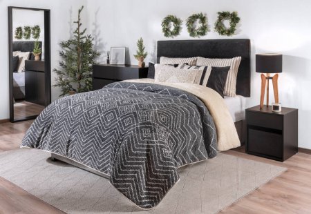 Photo for Modern Scandinavian Bedroom Interior with a Padded Fabric Headboard, Warm Black Cover, and Striped cozy Pillows on the Bed, beside a Wooden Nightstand Featuring Christmas Decorations. - Royalty Free Image