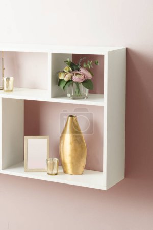 Photo for White Square Floating Bookshelf with Multi-Compartment Display, Featuring Gold Vase Ornaments, a Vase, and Picture Frame Mock-Up on a Light Pink Wall Background, in a Room Illuminated by Soft Light. - Royalty Free Image