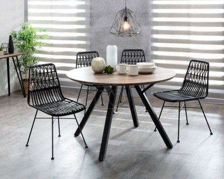 Dining Room Featuring a Mid-Century Modern Wooden Round Dining Table with Metal Legs, Patio Wicker Dining Chairs, White Tableware, Kitchen Utensils, and Windows with Zebra Blinds, Natural Light.