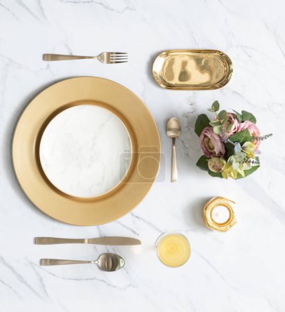 Event Celebration Wedding Dinner Table Setting with Shiny Wide Gold Rim Dinner Plate Set, Golden Cutlery, Gold Tableware, and Floral Decorations on White Marble Background, Flat lay Top View.