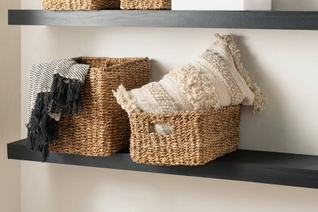Wicker Storage Baskets with Knitted Pattern Cotton Blankets and Textured Pillow on Wooden Floating Shelves Set in a Naturally Lit Living Room with Beige Wall.