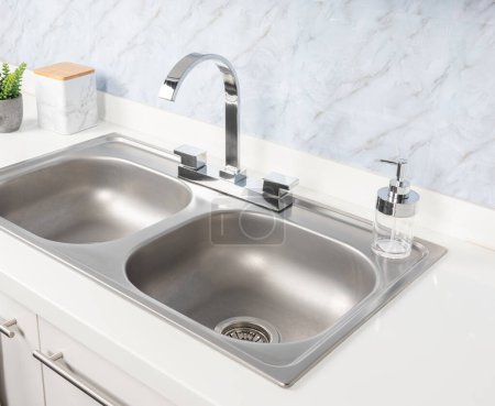 Close-Up of Double Basin Drop-In Stainless Steel Kitchen Sink and Faucet Mounted on a White Sink Base Cabinet in Bright Modern Kitchen, White Marble Background.