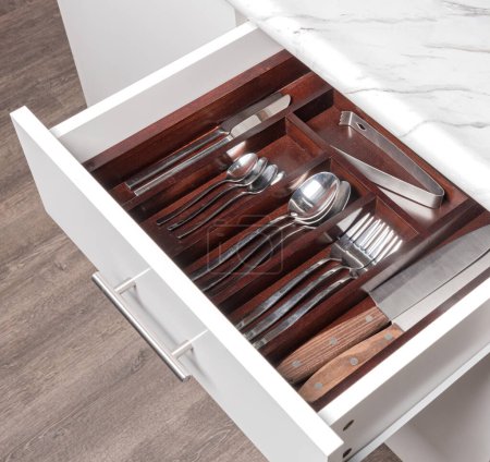 Top View of Open White Silverware Drawer with Wooden Dividers, Featuring Stainless Cutlery and White Marble Countertop, in Kitchen Cabinet over Natural Wooden Flooring.