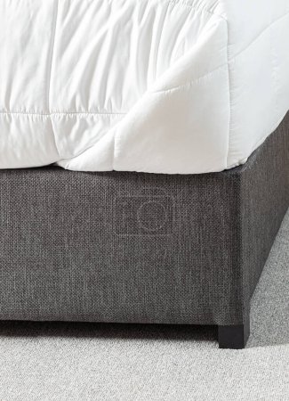 Nahaufnahme Detail Daunendecke, weiße Matratze Protector auf grauem niedrigen Stoff-Plattform-Bett im Schlafzimmer, über einem grauen Bereich Teppich.