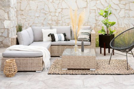 Photo for Spring Patio Furniture Set with Rattan Sectional Sofa, Decorative Pillow Covers, Wicker Patio Chairs, and Rattan Coffee Table, on a Botanical Area Rug in an Outdoor Living Space, Decorative Stone Wall - Royalty Free Image