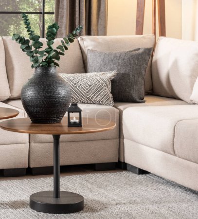 Photo for Living Room Featuring a Beige Fabric Upholstered Sectional Sofa with Decorative Throw Pillows and Mid-Century Modern Wooden Walnut Round Accent Coffee Table Set with Black Metal Legs, Ceramic Vase. - Royalty Free Image