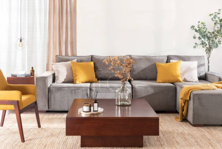 Photo for Modern Mid-Century Living Room Featuring a Large Grey Sectional Sofa with Chaise Lounge, Yellow Fabric Accent Armchair, Modern Wooden Square Storage Coffee Table, Area Rug, and Window with Curtains. - Royalty Free Image