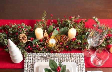 Photo for Elegantly set dinner table for Christmas with a festive red table runner, adorned garland with candles, pinecones, and red berries, beside a wine glasses and dinnerware, holiday dining ambiance. - Royalty Free Image