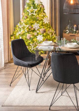 Photo for Elegant festive dining room with a lush Christmas tree adorned with golden ornaments, a glass-topped dining table set with white dishes and woven placemats, dark fabric chairs, beige area rug. - Royalty Free Image