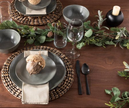 Elegant Christmas dinner setup on a wooden oak table showcasing gray plates set atop woven chargers, decorative wooden acorns on the plates, fresh greenery with pinecones, sleek black cutlery, napkins