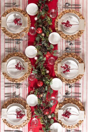 Photo for Festive Christmas dining table setup with elegant white plates adorned with bright red berry branches, set on woven placemats, amidst a vibrant red table runner, on a striped fabric backdrop, top view - Royalty Free Image