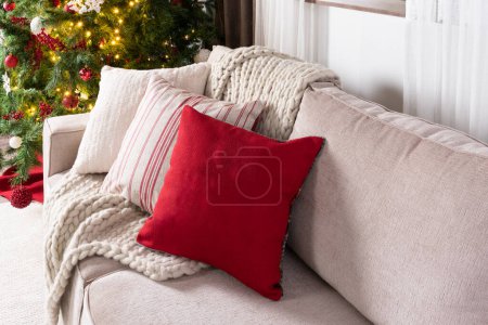 Gemütliches Weihnachtsambiente, neutrales Sofa mit strukturierten Kissen, rotes Akzentkissen, klobige Stricksachen, vor einem glitzernden Weihnachtsbaum, der von goldenen Lichtern erleuchtet wird, Großaufnahme