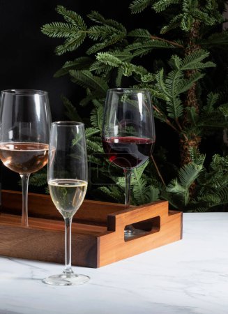 Foto de Elegante exhibición de copas de vino llenas de vino tinto y espumoso, en una bandeja de madera, ambientada en el fondo de un árbol de Navidad, ramas verdes, sobre una encimera de mármol blanco, fondo negro. - Imagen libre de derechos