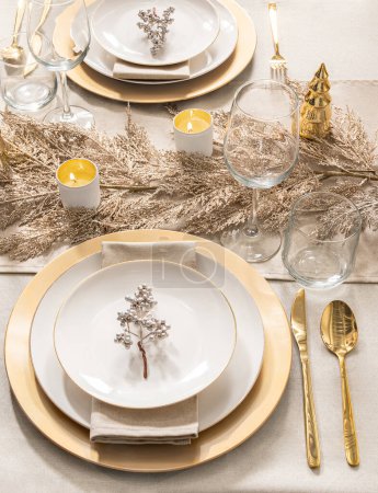 Table à manger élégante festive : Assiettes en porcelaine dorées scintillantes sur chargeurs dorés, associées à des couverts en or, des verres à vin en cristal et des bougies en ivoire au milieu d'un décor de feuillage doré, vue de dessus.
