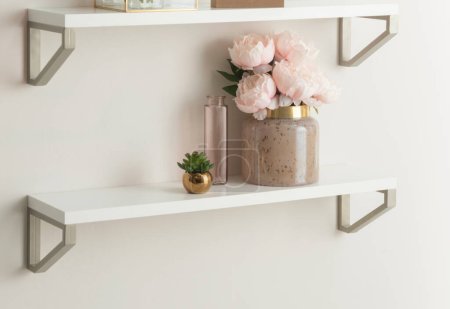 Photo for Elegant Floating Shelves Display: A Chic Ceramic Vase with Blush Pink Peonies, a Petite Glass Bottle, and a Small Gold Pot with a Succulent, Creating a Serene and Stylish Wall Accent in a Modern Home. - Royalty Free Image