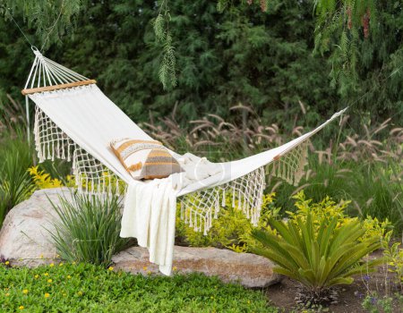 Photo for Tranquil Garden Oasis Featuring a White Cotton Hammock with Fringe Detail, Adorned with a Striped Cushion and Knit Throw, Nestled Among Lush Greenery and Vibrant Yellow Flowers, with a Majestic Fern. - Royalty Free Image