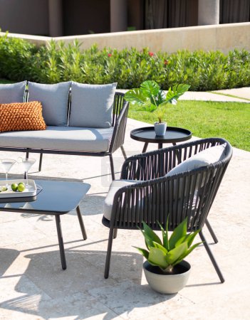 Foto de Moderno espacio de relajación al aire libre con un elegante sofá de Wireframe negro y sillones a juego con cojines grises, una mesa de café redonda, una planta de serpiente en maceta ceñida, en una terraza en medio de arbustos verdes. - Imagen libre de derechos