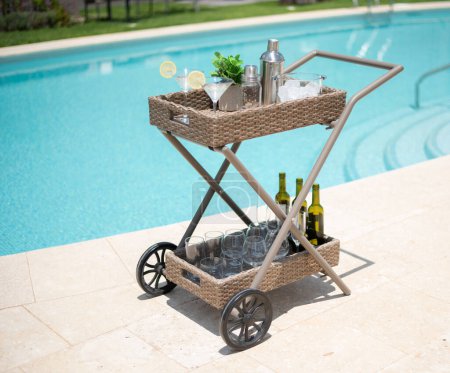 Panier de célébration au bord de la piscine d'été équipé d'un chariot à roulements de chariot en rotin, d'un agitateur en acier inoxydable et d'un rayon de bouteilles de vin blanc, préparé pour une fête en plein air sur des tuiles ensoleillées.