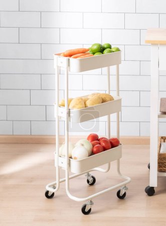 Photo for Modern Kitchen Organization with a Three-Tier White Metal Rolling Storage Cart Utility Organizer Laden with Vegetables, Carrots, Limes, Potatoes, and Tomatoes, in a Contemporary Kitchen Setting - Royalty Free Image