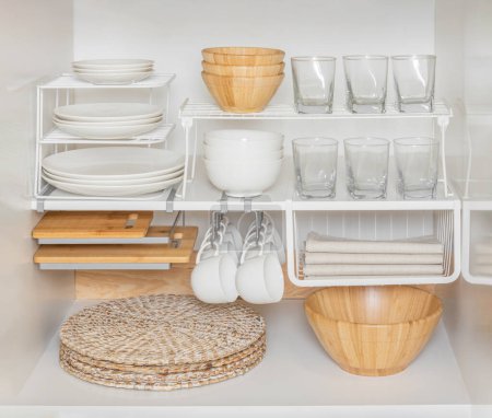 Organización óptima del gabinete de cocina Mostrando una matriz de vajilla: placas de porcelana blanca, vasos de vidrio, tableros de corte, tazones de cerámica, manteles y servilletas de lino en un espacio culinario brillante.