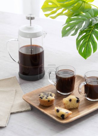 Stärkendes Frühstück am Morgen: Französische Presse-Kaffeemaschine gefüllt mit reichhaltigem dunklen Kaffee, gepaart mit Glasbechern und frisch gebackenen Blaubeer-Muffins auf einem hölzernen Serviertablett, bereit für den Tag