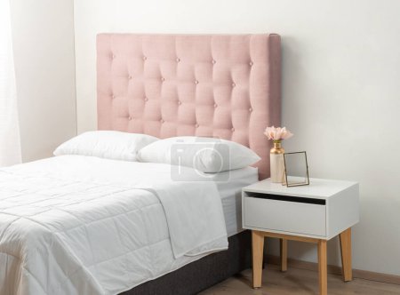 Téléchargez les photos : Chic Feminine Bedroom : Plush Pink Tufted Headboard Setting the Tone for a Delicate Space, complétée par une table de chevet blanche contemporaine avec des jambes en bois, ornée d'une fleur de pastel dans un vase en or. - en image libre de droit