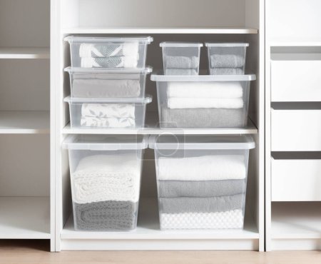 Contemporary white closet interior with transparent plastic sweater storage boxes organizing folded towels and soft blankets, complementing the clean, organized bedroom ambiance, minimalist design.