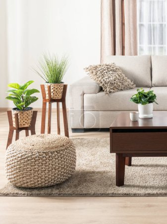 Warmly Lit Contemporary Living Room, Beige Textured Sofa with a Shaggy Pillow, a Woven Round Ottoman, Wooden Plant Stands with Greenery, a Modern Coffee Table, Upon a Beige Rug Over wooden Flooring.