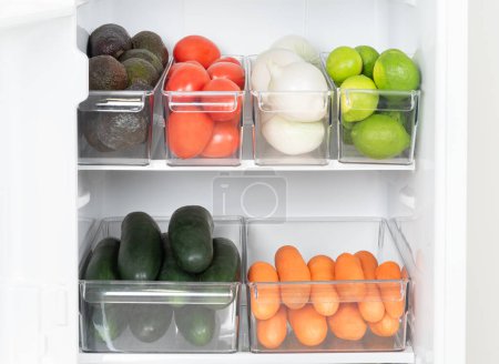 Réfrigérateur intérieur bien organisé doté de bacs en plastique transparents soigneusement garnis d'un assortiment de fruits et légumes sains : avocats, tomates, oignons blancs, chaux, planification des repas sains.