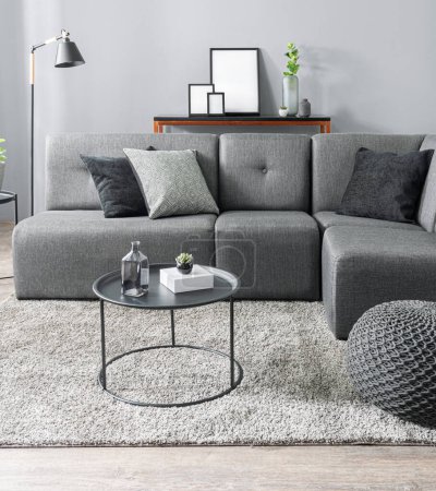 Modernes monochromes Wohnzimmer mit einem anthrazitgrauen modularen Sofa, geschmückt mit geometrischen und samtenen Kissen, einem runden Couchtisch aus Metall, einem strukturierten Hocker auf einem zotteligen beigen Teppich, schlanken Konsolentischen mit Akzenten