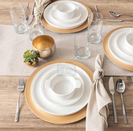 Sophisticated Table Setting with Gold-Rimmed White Porcelain Plates, Elegant Glassware, Stainless Steel Flatware, Linen Napkins with Knot Detailing, on a Natural Textured Table Runner, Top View.