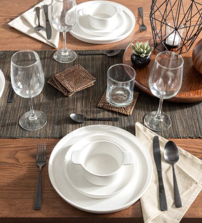 Elegant Dining Table Setting Featuring Pristine White Porcelain Dishware, Crystal Stemware, and Black Flatware, Arranged on a Textured Striped Table Runner, Woven Coasters and a Decorative Objects.