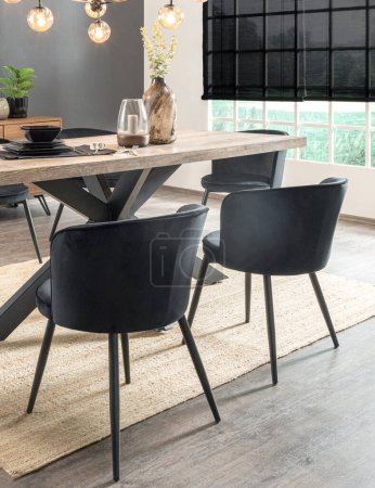 Modern Dining Room with Industrial Flair, Robust Wooden Table with Crossed Metal Legs, Elegant Black Upholstered Chairs, Under a Edison Bulb Lighting, Accented by a Metallic Vase and Black Tableware.