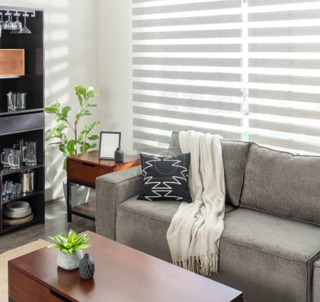 Contemporary Apartment Living Room with a Grey Sofa, Patterned Throw Pillow and Cream Blanket, a Sleek Bar Cabinet with Glassware and Wine Essentials, Illuminated by Natural Light, Striped Curtains.
