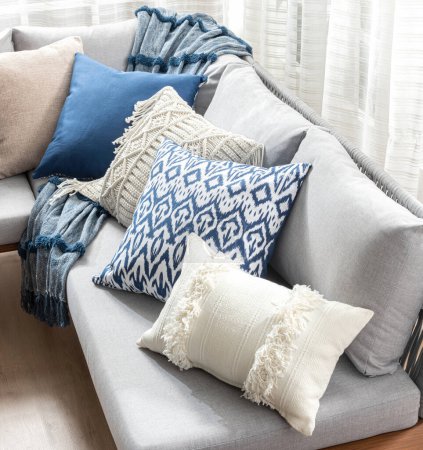 Acogedor rincón de la sala de estar con un sofá gris suave adornado con una matriz de cojines decorativos varias texturas y patrones, incluyendo azul sólido, beige y una manta de tiro, contra cortinas que fluyen.