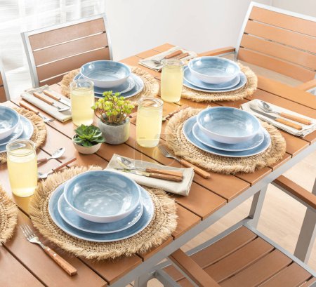 Frühlings-Sommer-Speiseplan mit himmelblauen Keramiktellern auf natürlich gewebten Tischsets, hölzernem Besteck, erfrischender Limonade in klaren Gläsern, auf einem warmen Holzlattentisch mit sonnigem Ambiente.