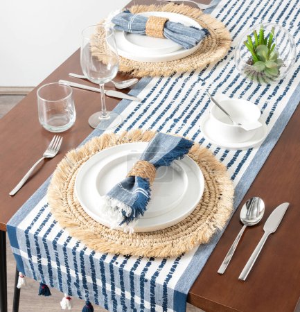 Mediterran inspirierte sommerliche Tischdekoration mit knackigem weißem Porzellan auf natürlichen gewebten Raffia-Tischsets, mit kühnem blau gestreiftem Leinen-Tischläufer und passenden Servietten, klarem Glasbesteck.