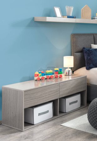Dormitorio moderno para niños con pared de acento azul con una elegante unidad de almacenamiento gris con contenedores de tela y tapa de madera que muestra un colorido conjunto de trenes de madera, iluminación suave y elementos decorativos.